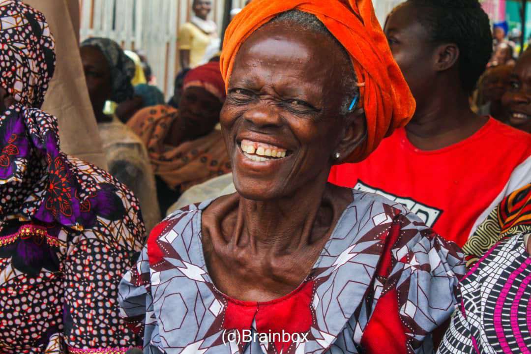 An elderly woman in a red and orange headwrap smiles broadly at the outreach