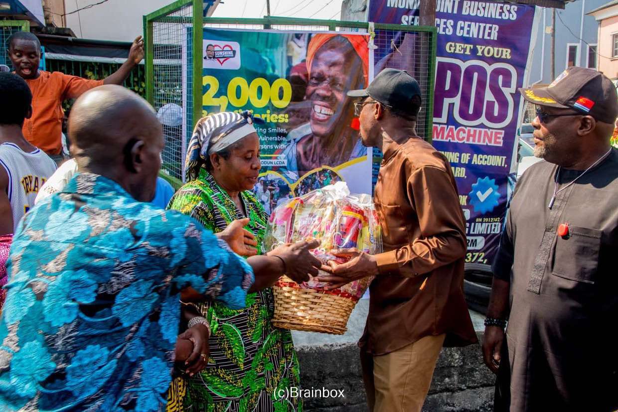 Dr. Arogundade Samsondeen hands a festive basket to an elderly woman beside the 2,000 beneficiaries banner
